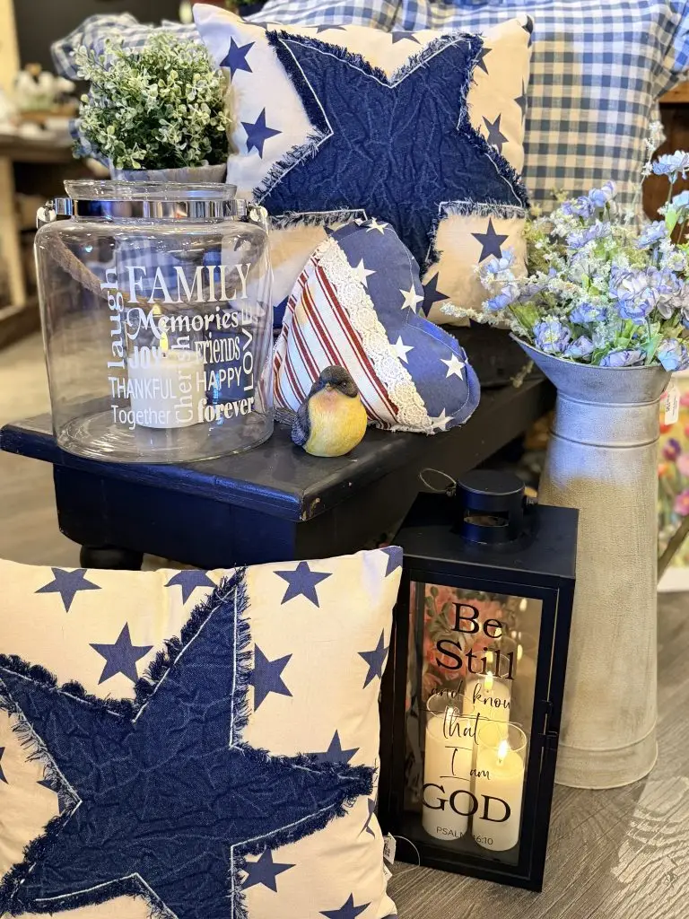 Patriotic farmhouse-style home décor display featuring blue star pillows, an American flag heart pillow, faux blue flowers in a metal vase, a decorative lantern with candle, and a glass jar with inspirational family quote.