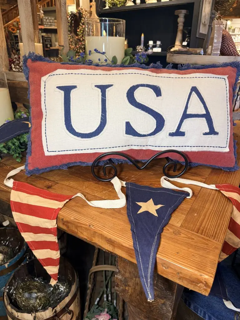 Rustic patriotic décor display featuring a red, white, and blue “USA” pillow with vintage-style bunting on a wooden table in a farmhouse gift shop.